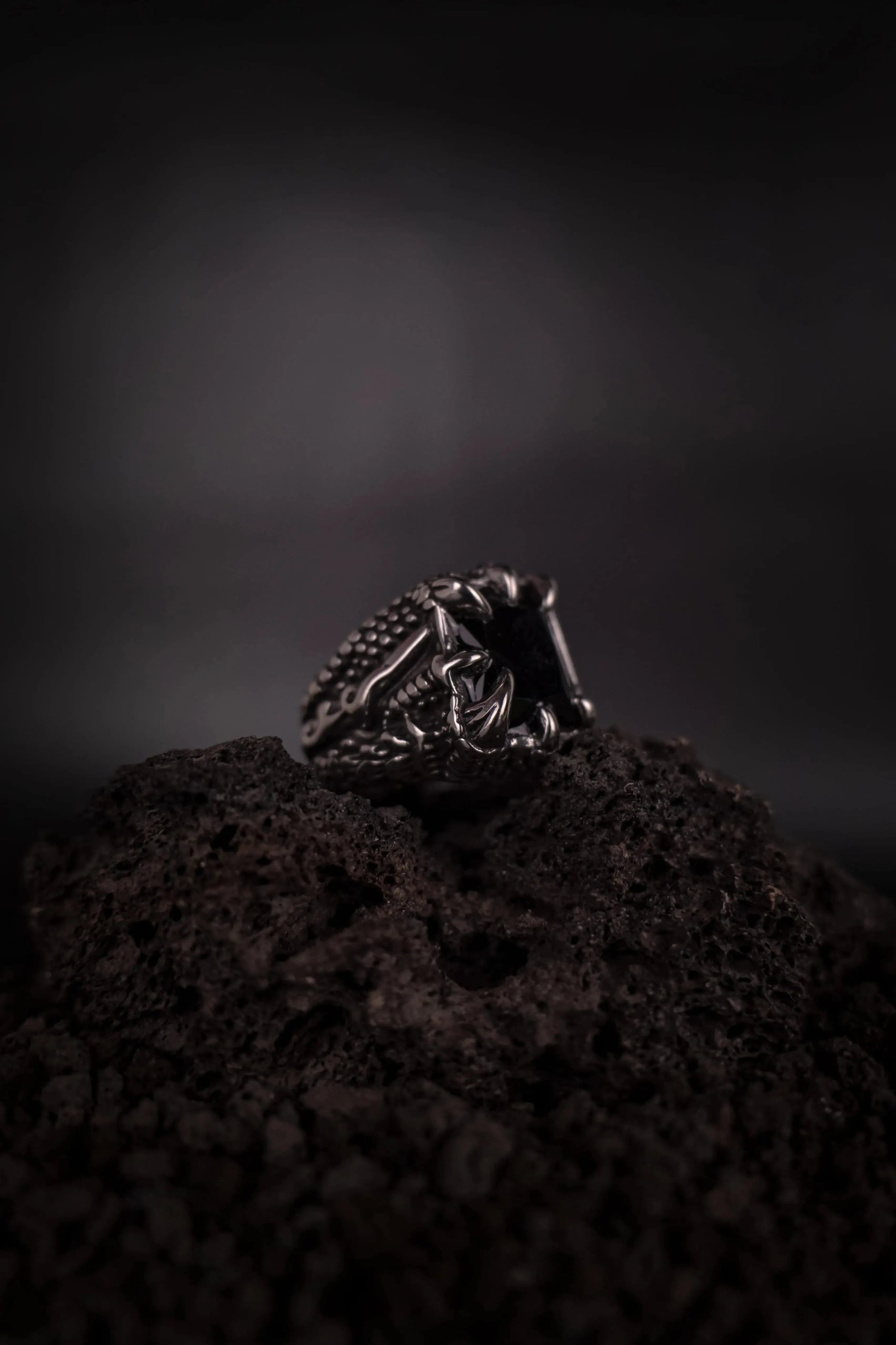bague chevalière style biker rebel rock avec une pierre noir tenue par des griffes posé sur de la roche volcanique