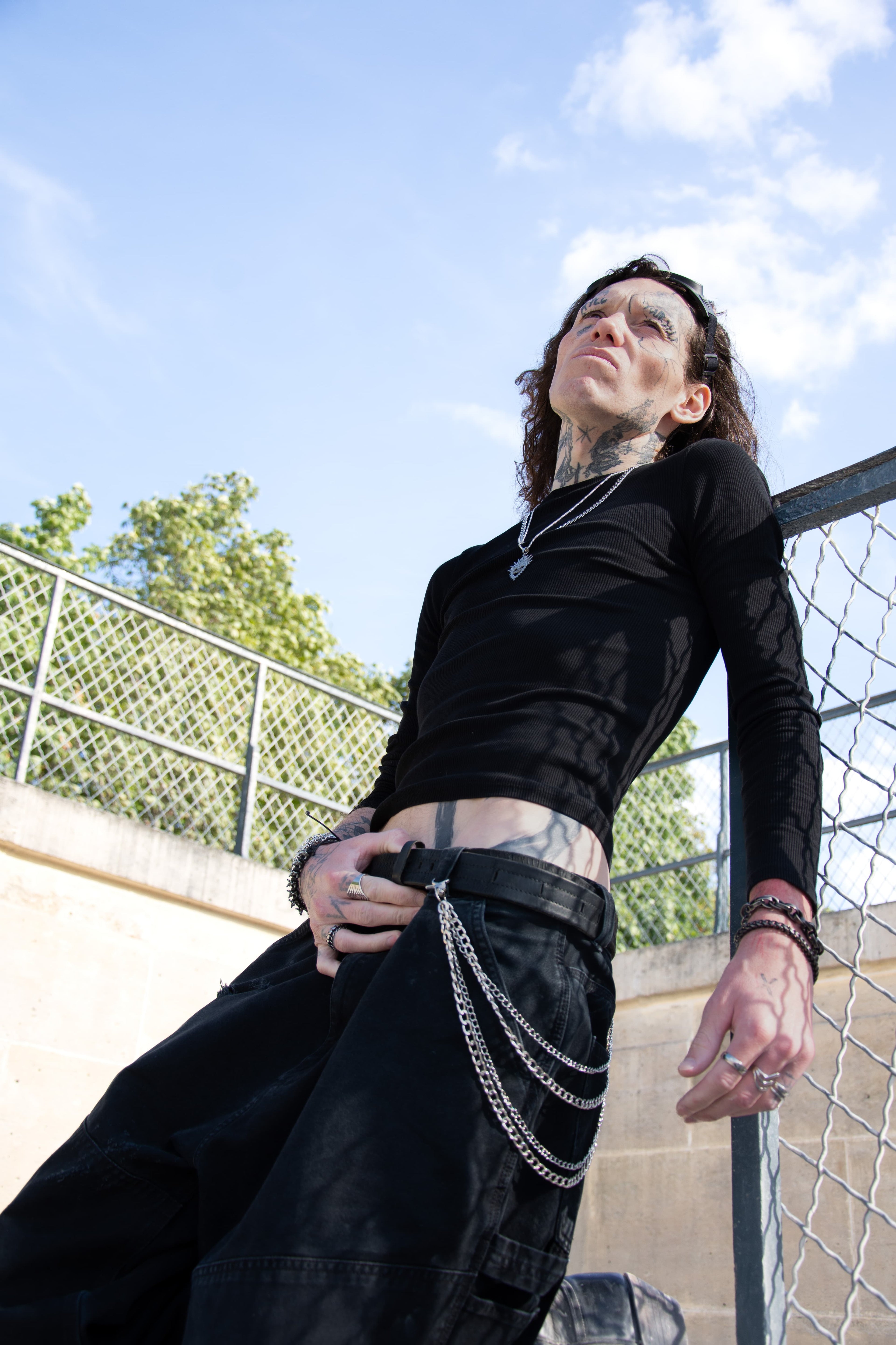 Mannequin tatoué posant contre une grille en portant les chaines de pantalon sur une tenue full black.