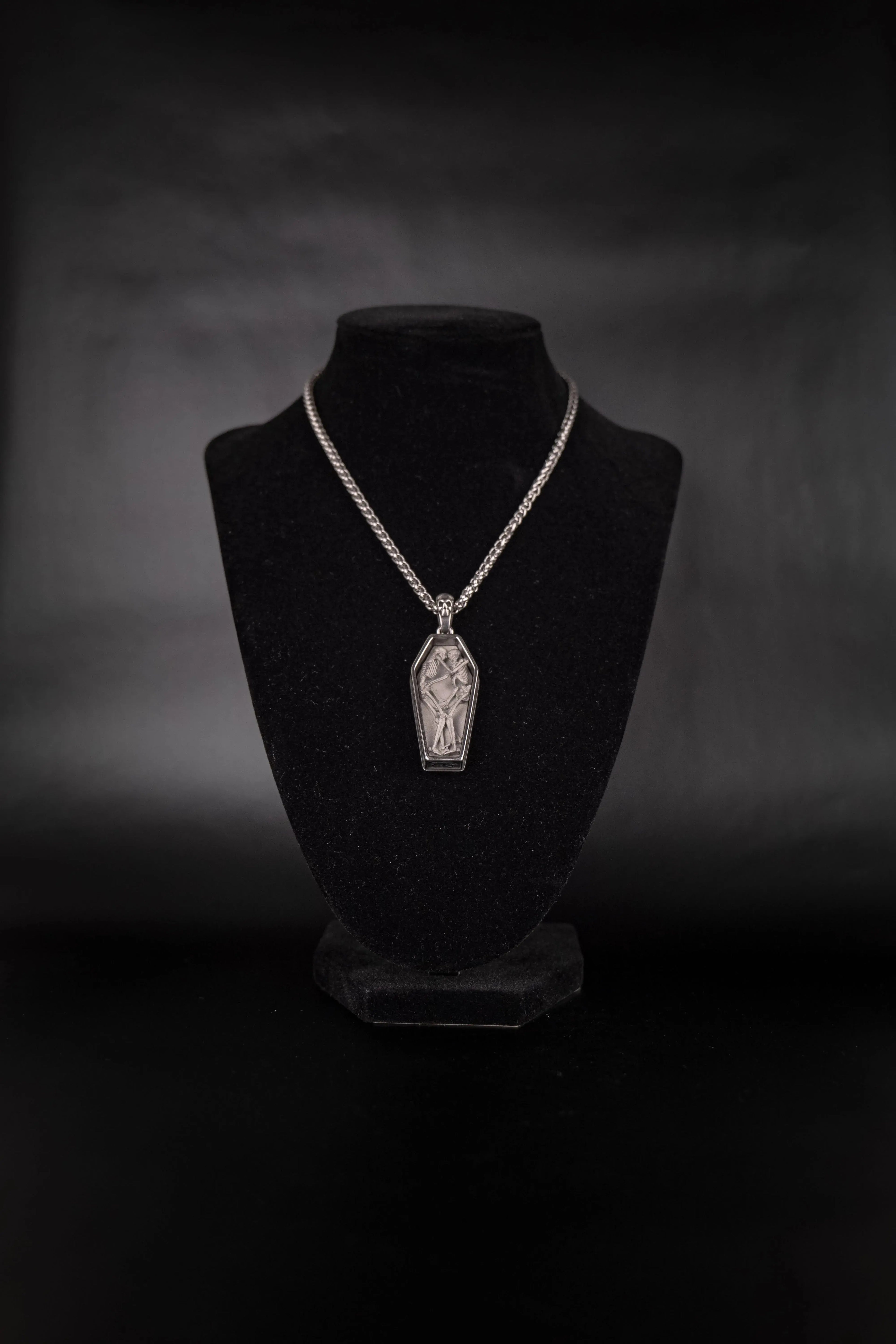 Pendentif cercueil contenant deux squelette amoureux, enlacé l'un à l'autre porté sur un présentoir noir.