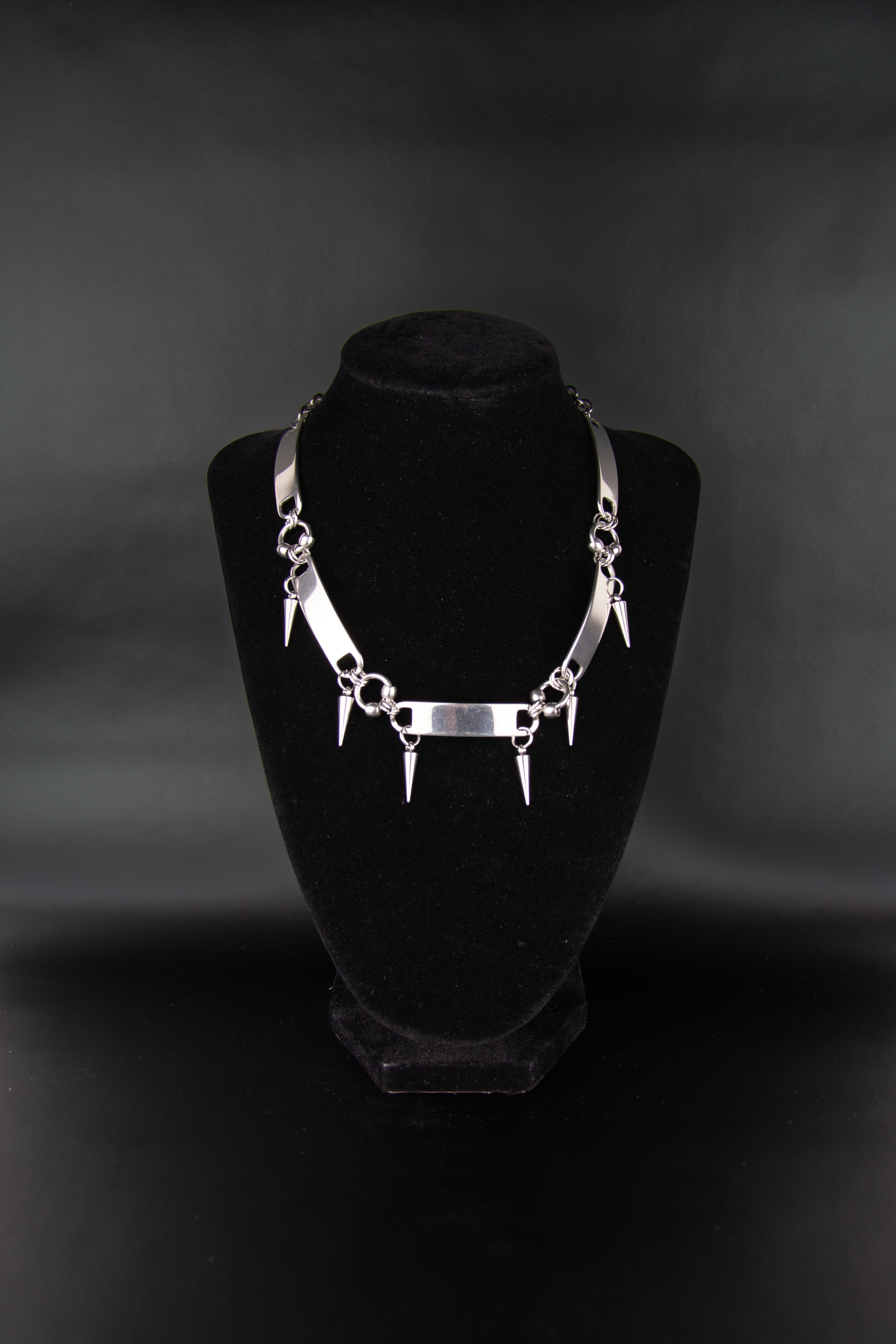 Collier ras de cou avec piques et métallique tab pour lacet de chaussure posé sur un présentoir à bijoux en velours noir et fond noir. 