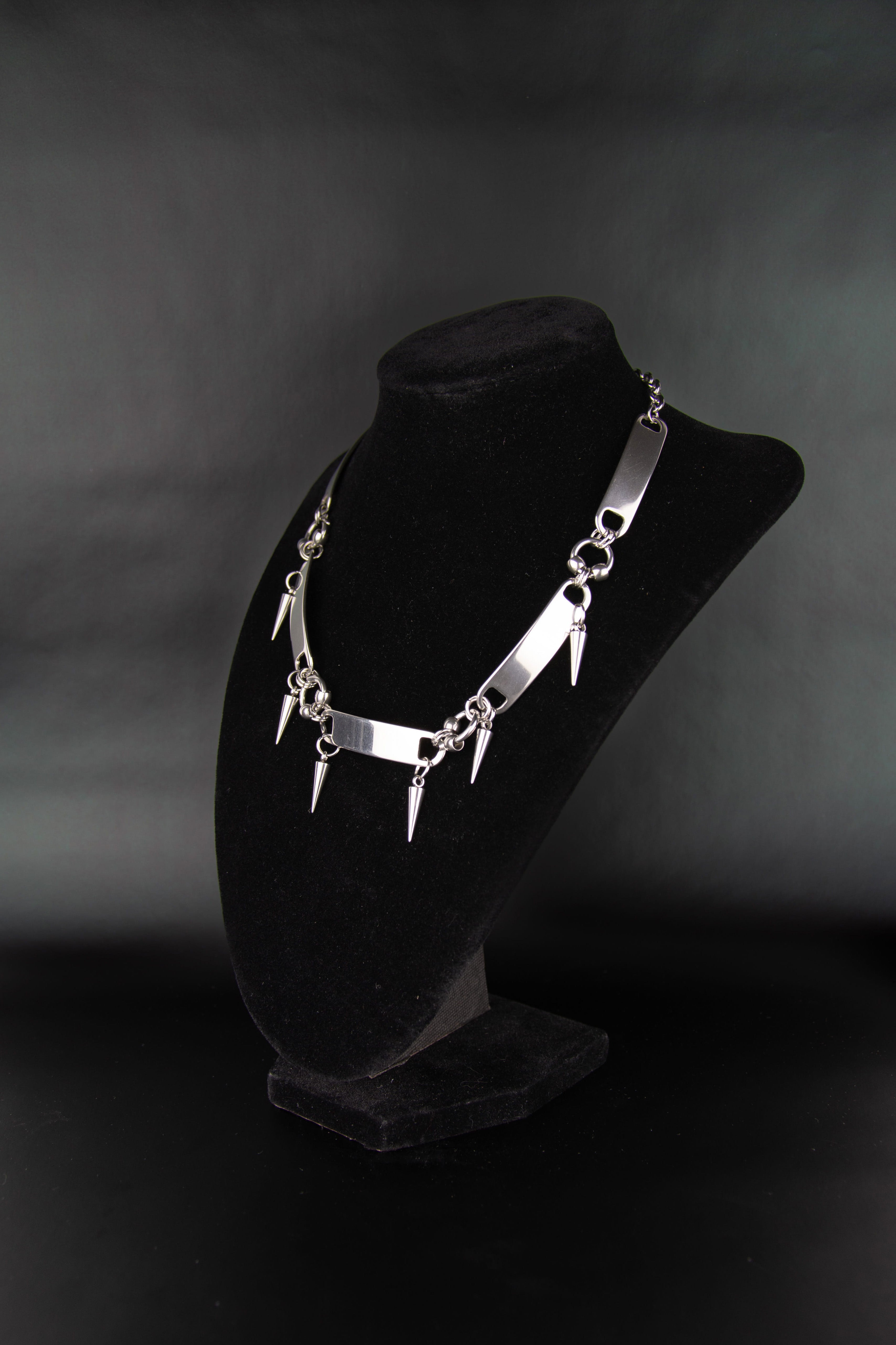 Collier ras de cou avec piques et métallique tab pour lacet de chaussure posé sur un présentoir à bijoux en velours noir de trois quarts et fond noir. 