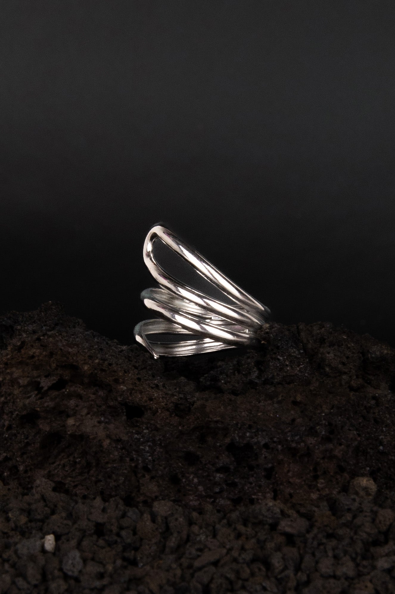 Bague asymétrique ouverte et réglable en acier inoxydable posée de trois quarts sur une roche volcanique noir.